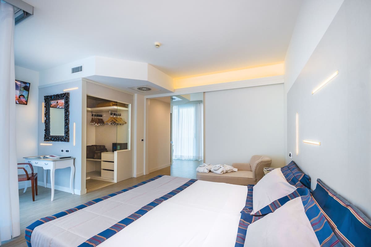 A modern hotel room with a large bed, a desk, a chair, a wardrobe, and a seating area.