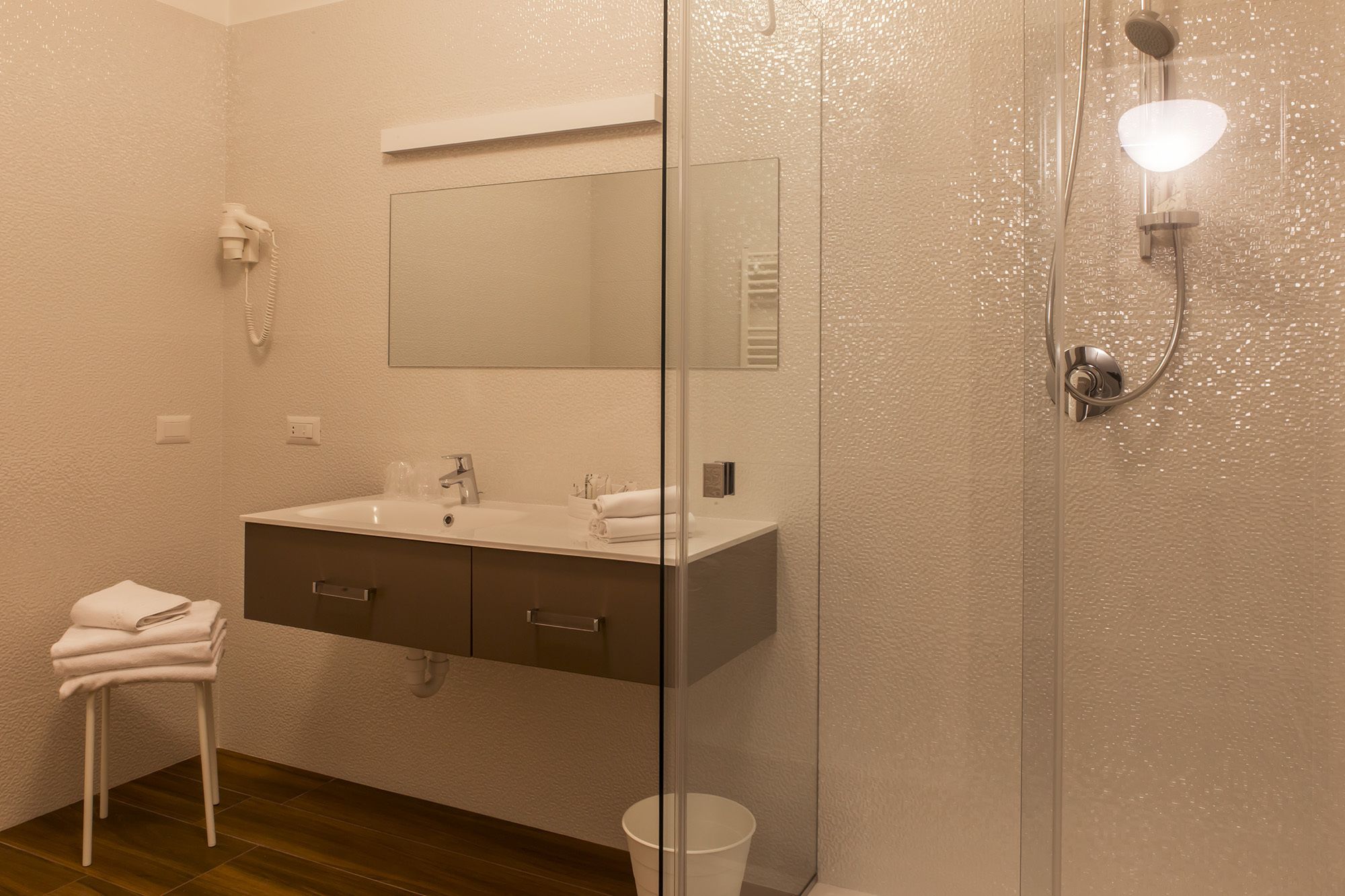 A bathroom with a white sink, mirror, and shower. There is a towel rack and a white bucket.