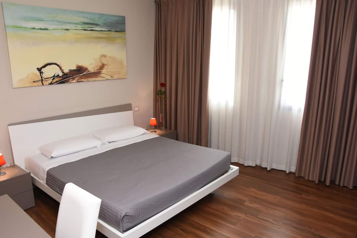 A neatly made bed with a grey bedspread, a white pillow, and a headboard. There is a nightstand with a red lamp and a vase with a single red rose. A painting of a beach scene is on the wall above the bed.