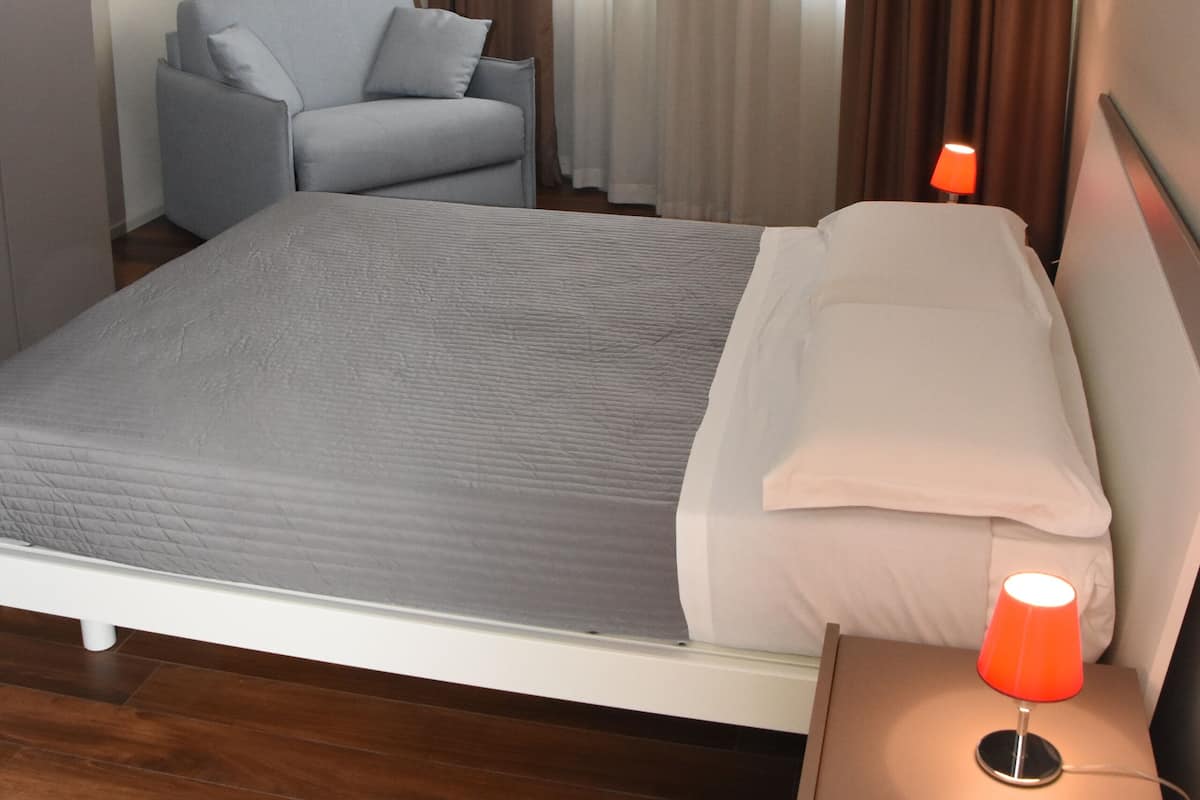 A hotel room with a large bed, a chair, a nightstand with a lamp, and a window with curtains.