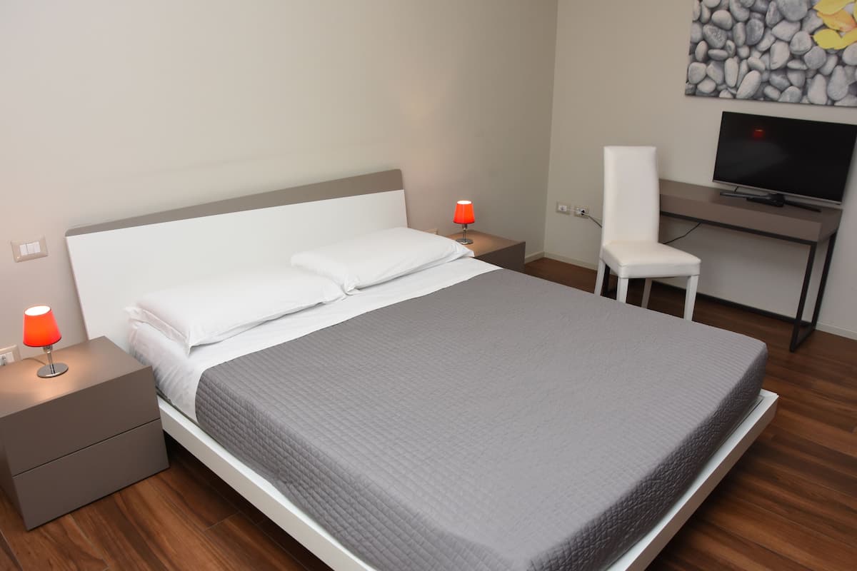 A neatly made bed with a grey quilt, two bedside tables with red lamps, a white chair, and a TV on a desk.