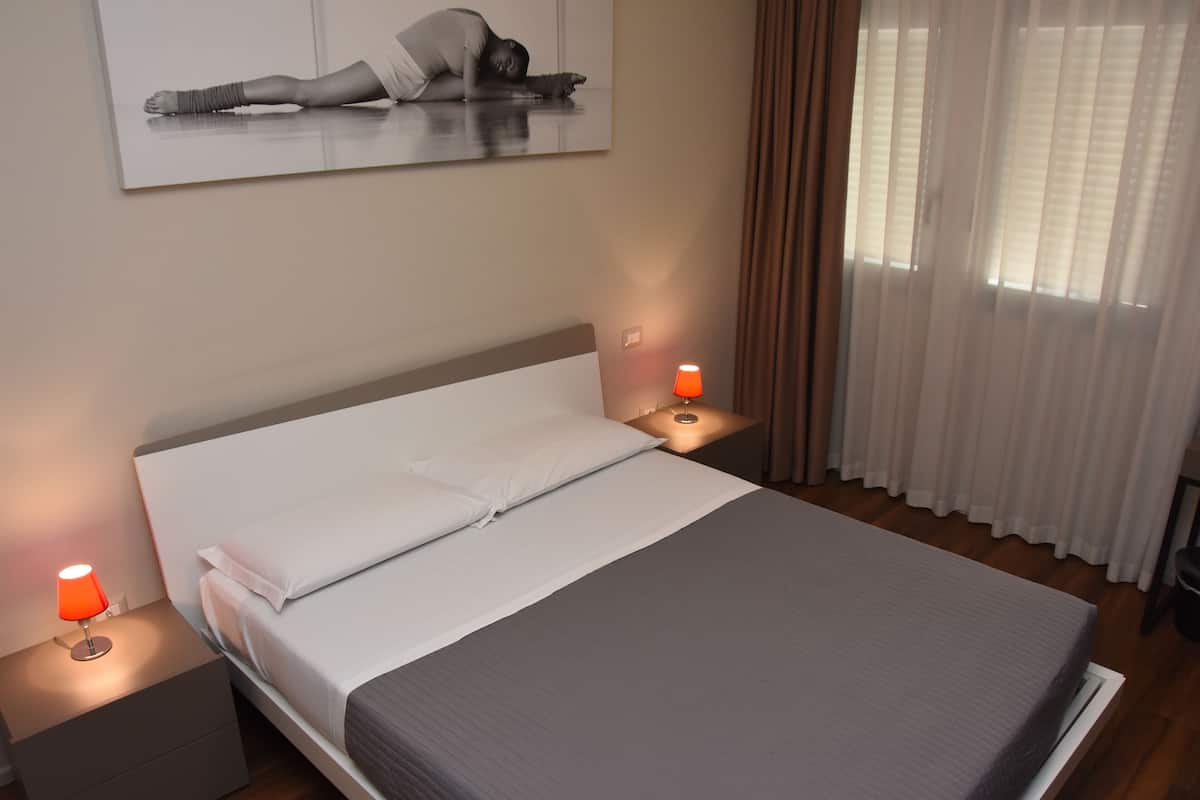 A neatly made bed with white and gray linens, two bedside lamps, a nightstand, and a painting on the wall.