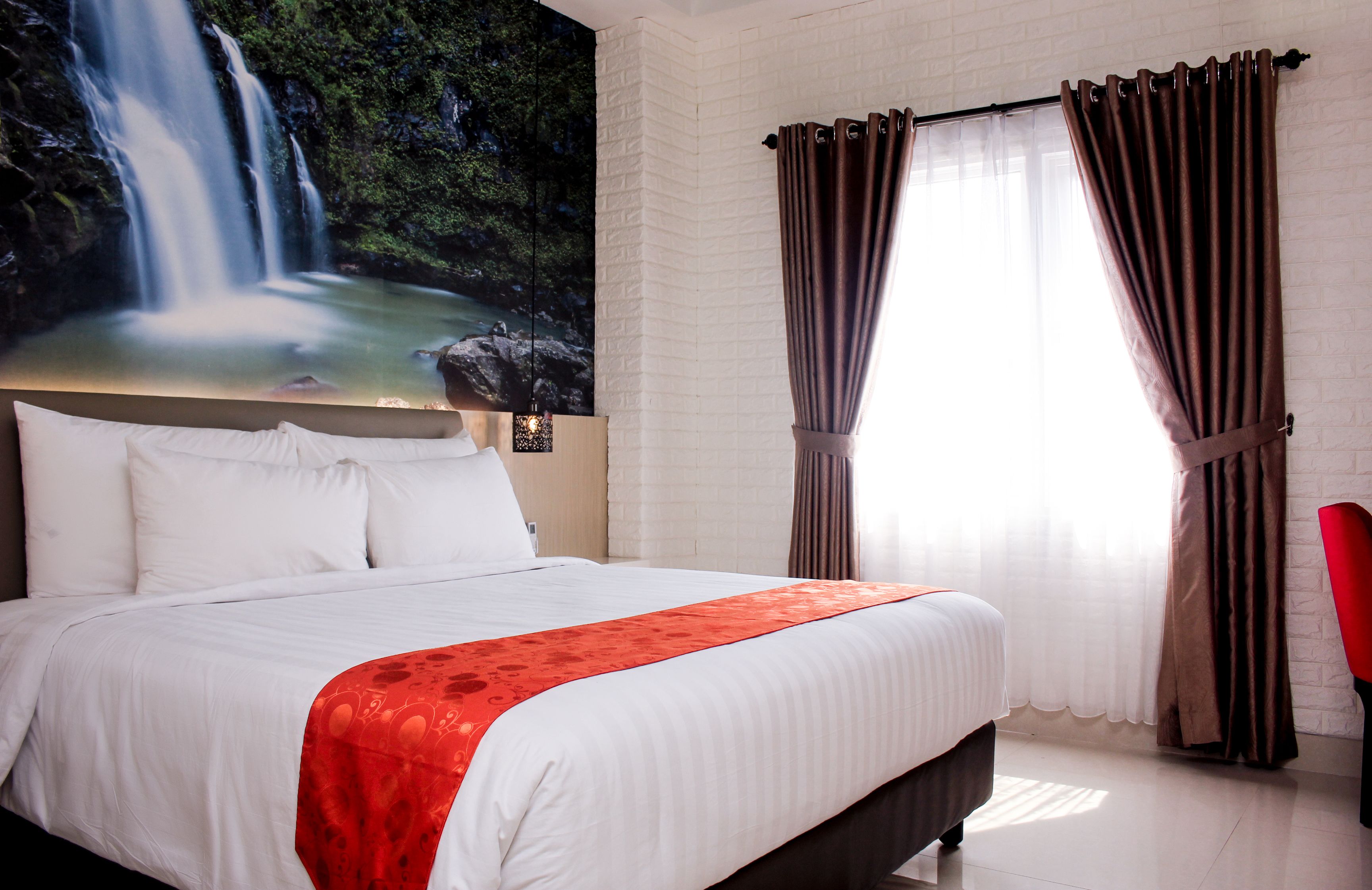 A hotel room with a large bed, white bedding, a red throw, a mural of a waterfall, and brown curtains.