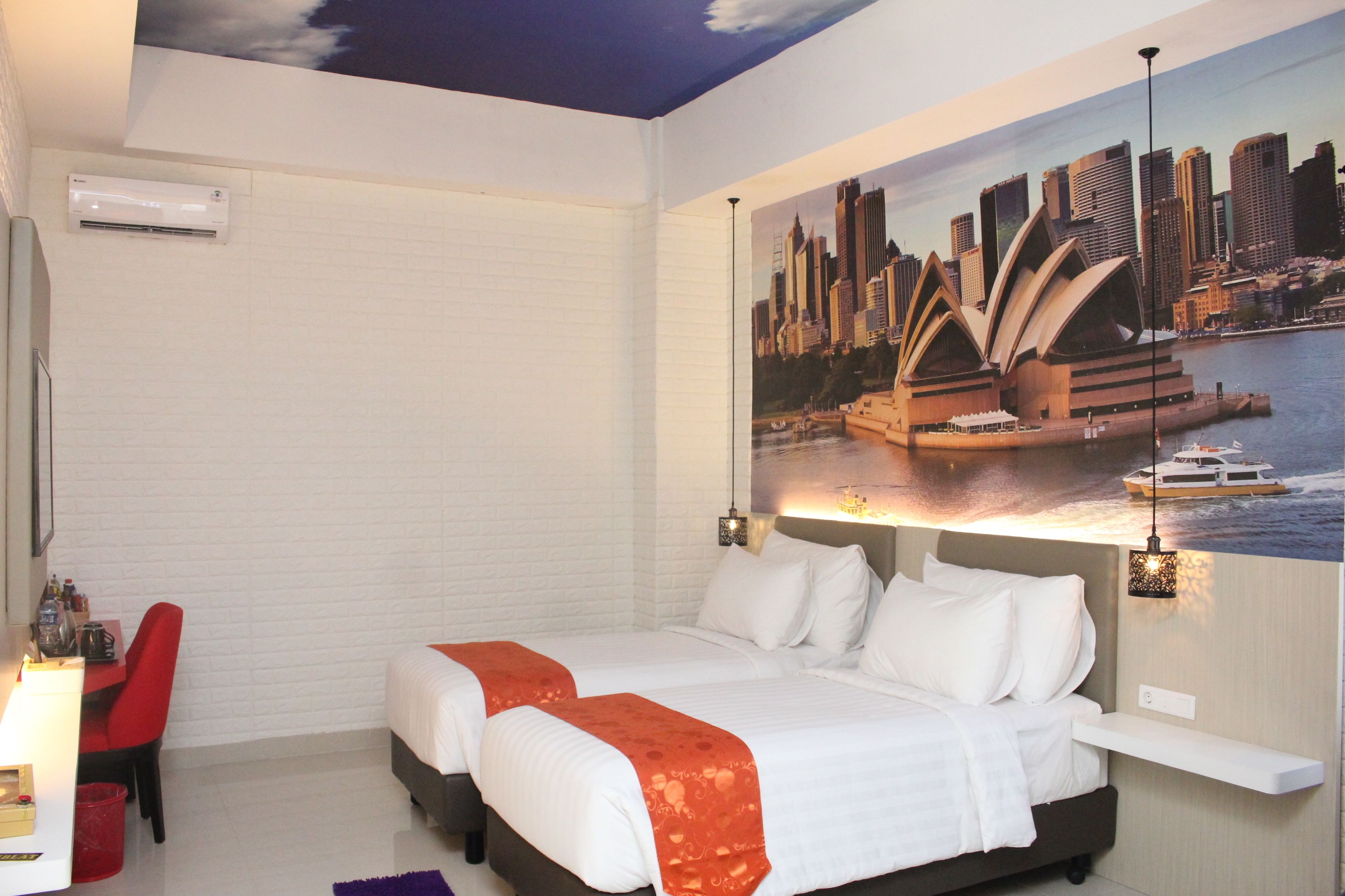 A hotel room with two beds, a red chair, a desk with a lamp, and a cityscape mural on the wall.
