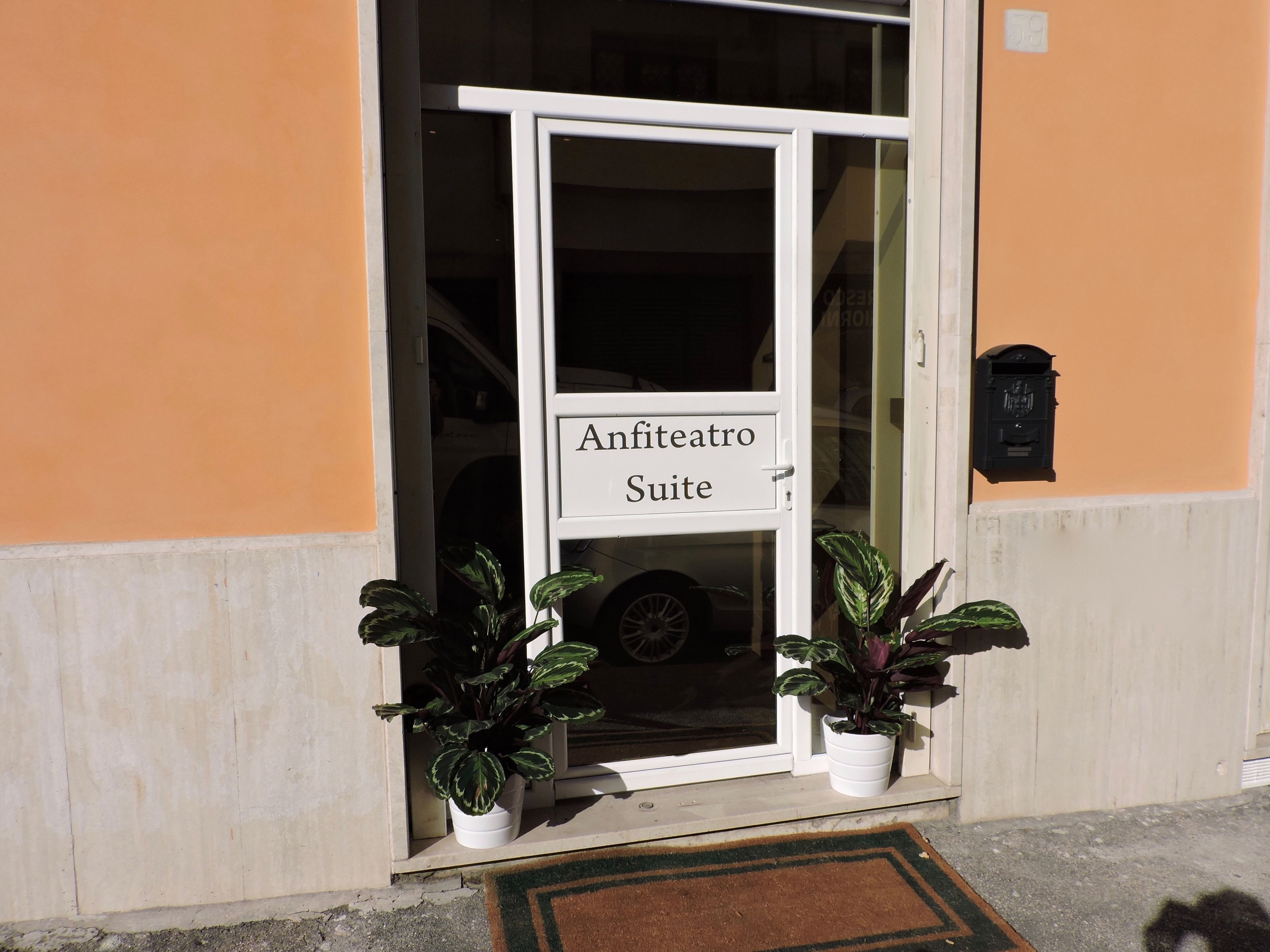 Entrada de hotel con un letrero que dice "Anfiteatro Suite". Hay dos plantas en macetas a cada lado de la puerta.