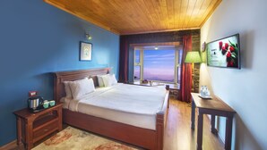 Habitación de hotel con una cama grande, cabecero de madera, una mesita de noche con hervidor, una mesa con lámpara y vistas al atardecer a través de una ventana.