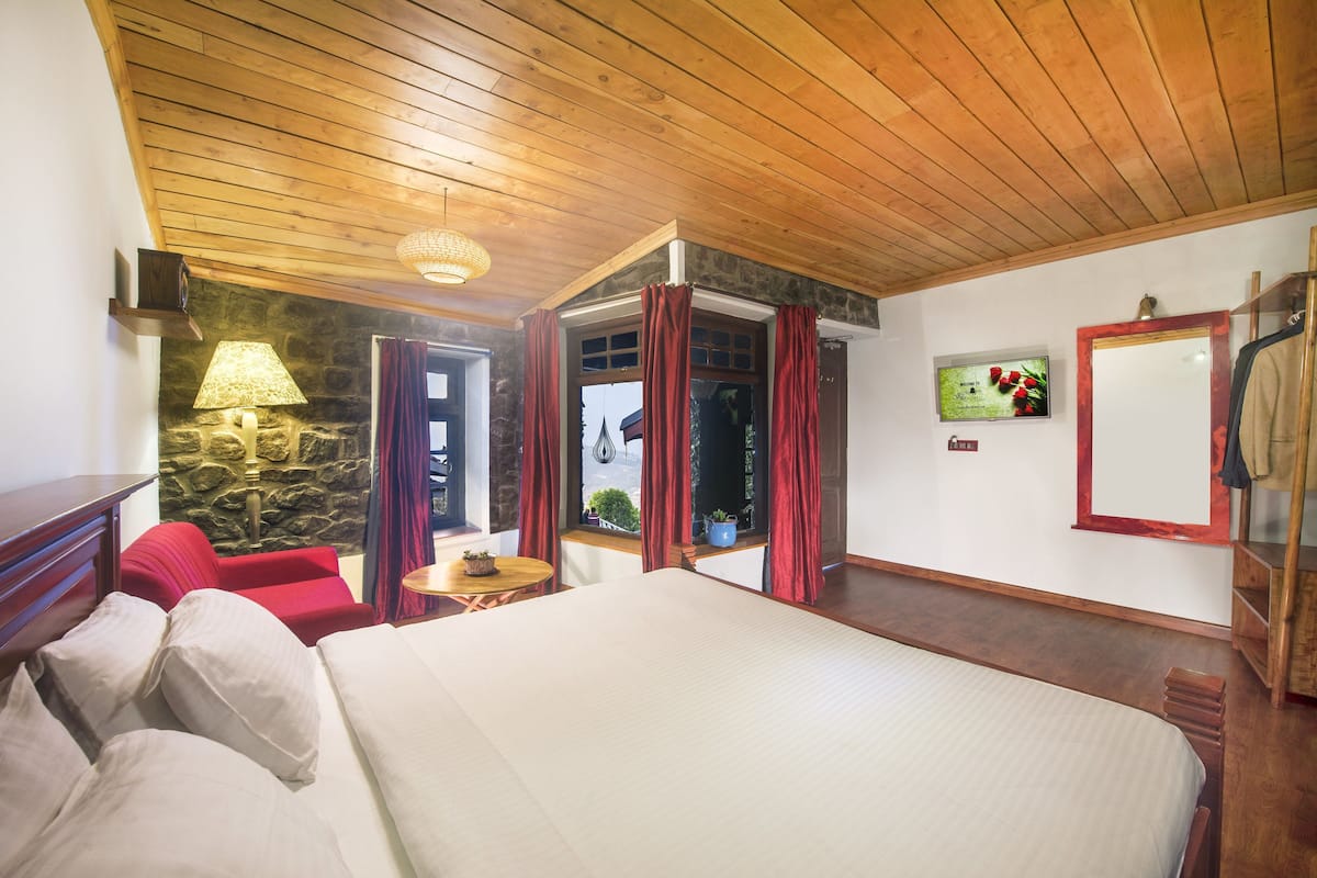 Une chambre à coucher avec un lit, un fauteuil rouge, une lampe, un mur en pierre, une fenêtre avec des rideaux et un miroir.