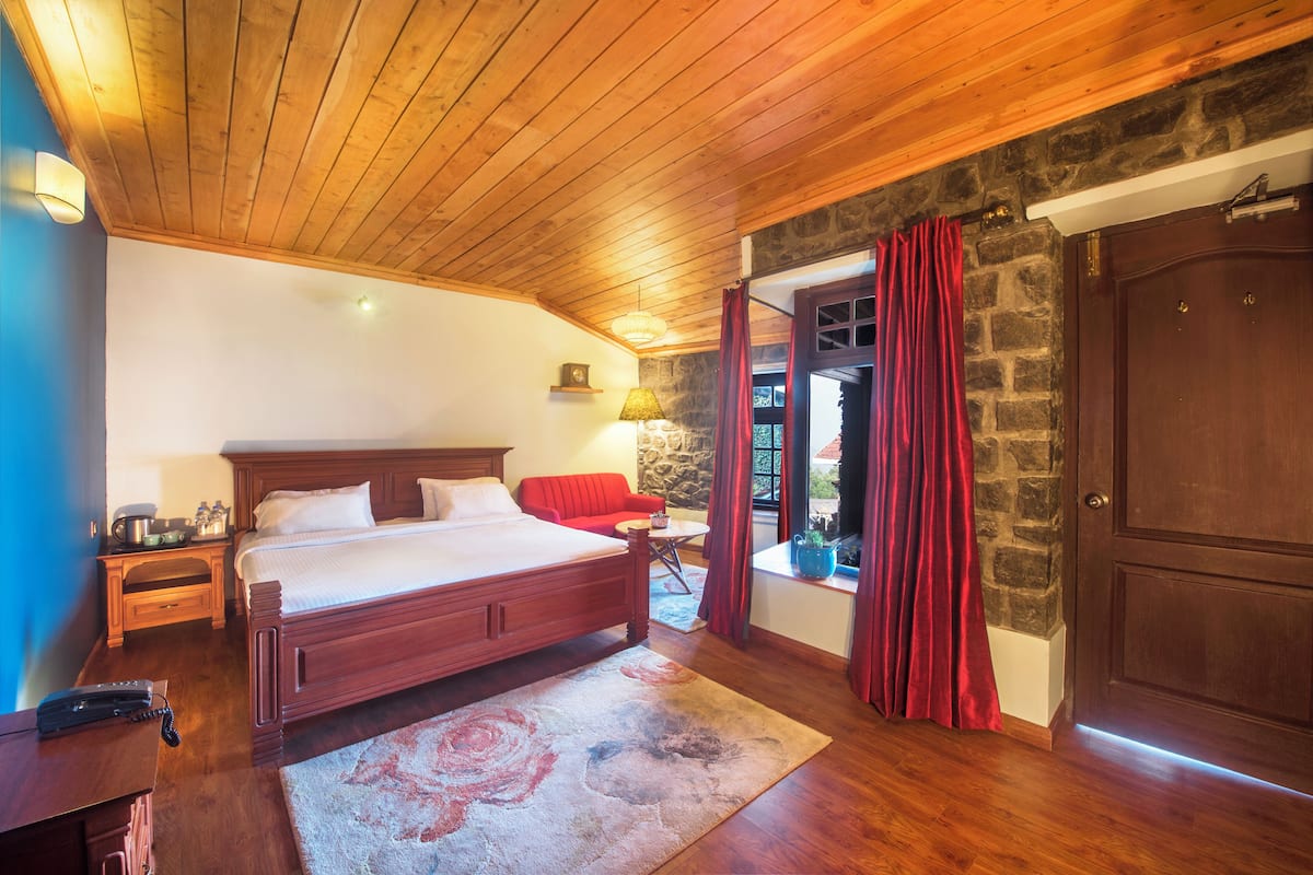 Une chambre avec un plafond en bois, un mur en pierre, un lit, un canapé rouge, une petite table avec une lampe et une porte donnant sur un espace extérieur.