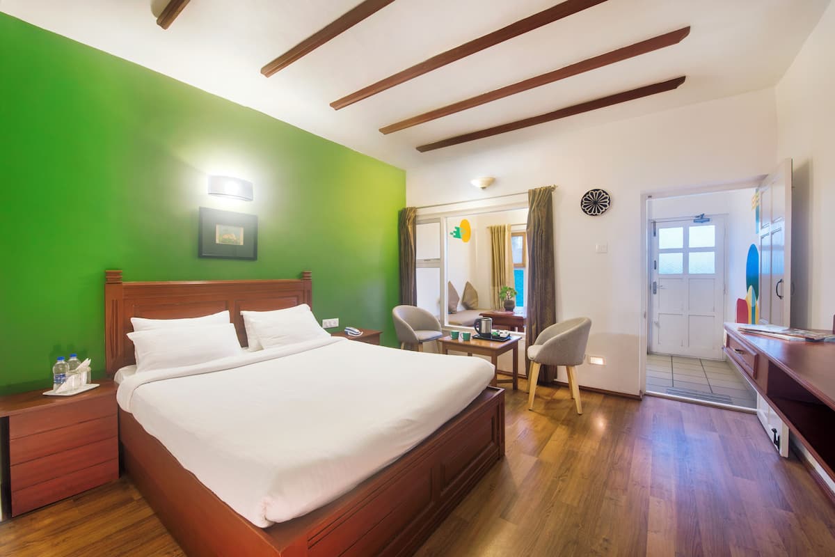 Une chambre d’hôtel avec un grand lit, une tête de lit en bois, des murs verts, un bureau avec une chaise et une porte donnant sur une autre pièce.