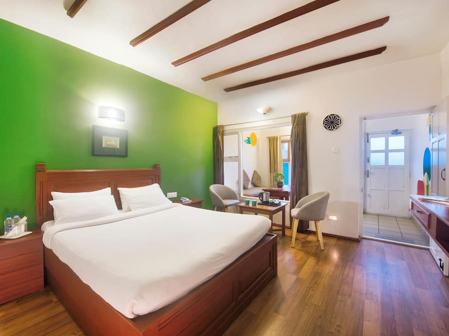 A hotel room with a large bed, wooden headboard, green walls, a desk with a chair, and a door leading to another room.