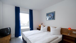 A hotel room with a large bed, a TV on a wooden table, a nightstand with a lamp, and a window with blue curtains. - Haus Nordstrand (Göhren)