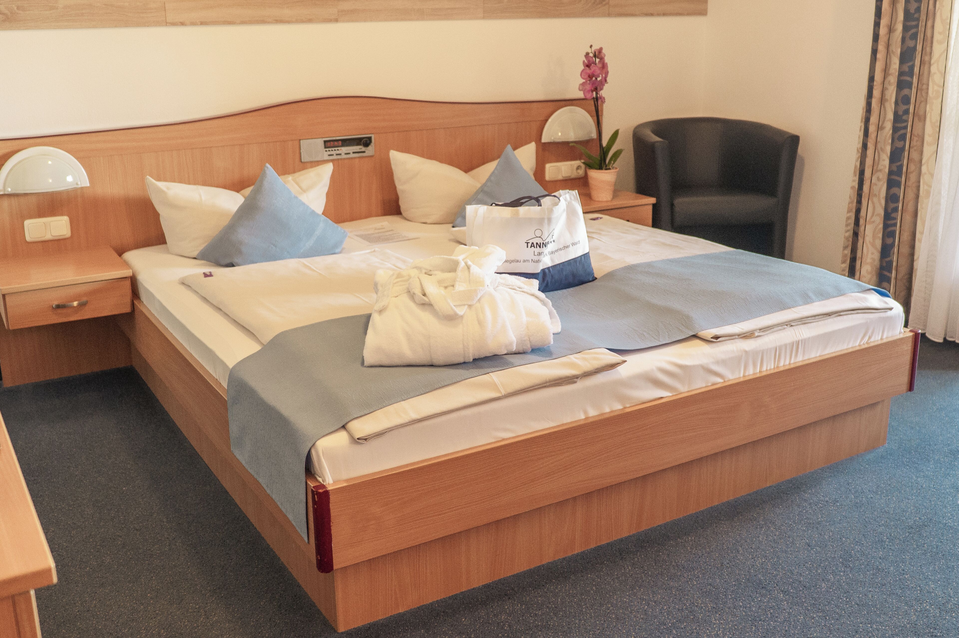Habitación de hotel con cama de madera, almohadas, mesita de noche con lámpara, una silla y una planta en maceta.