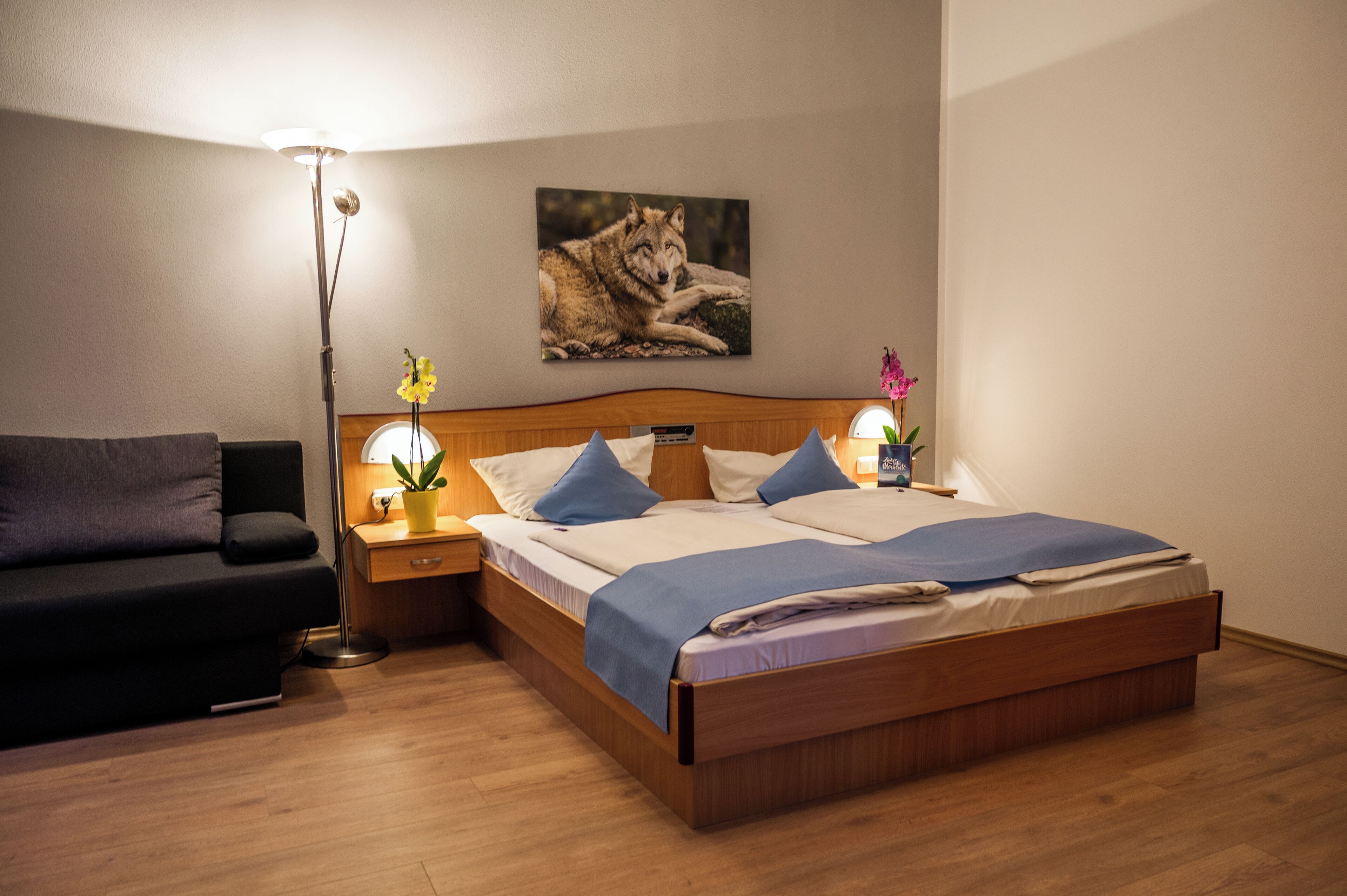 A neatly arranged bedroom with a wooden bed, a nightstand with a lamp, a wall-mounted picture of a wolf, and a potted plant.