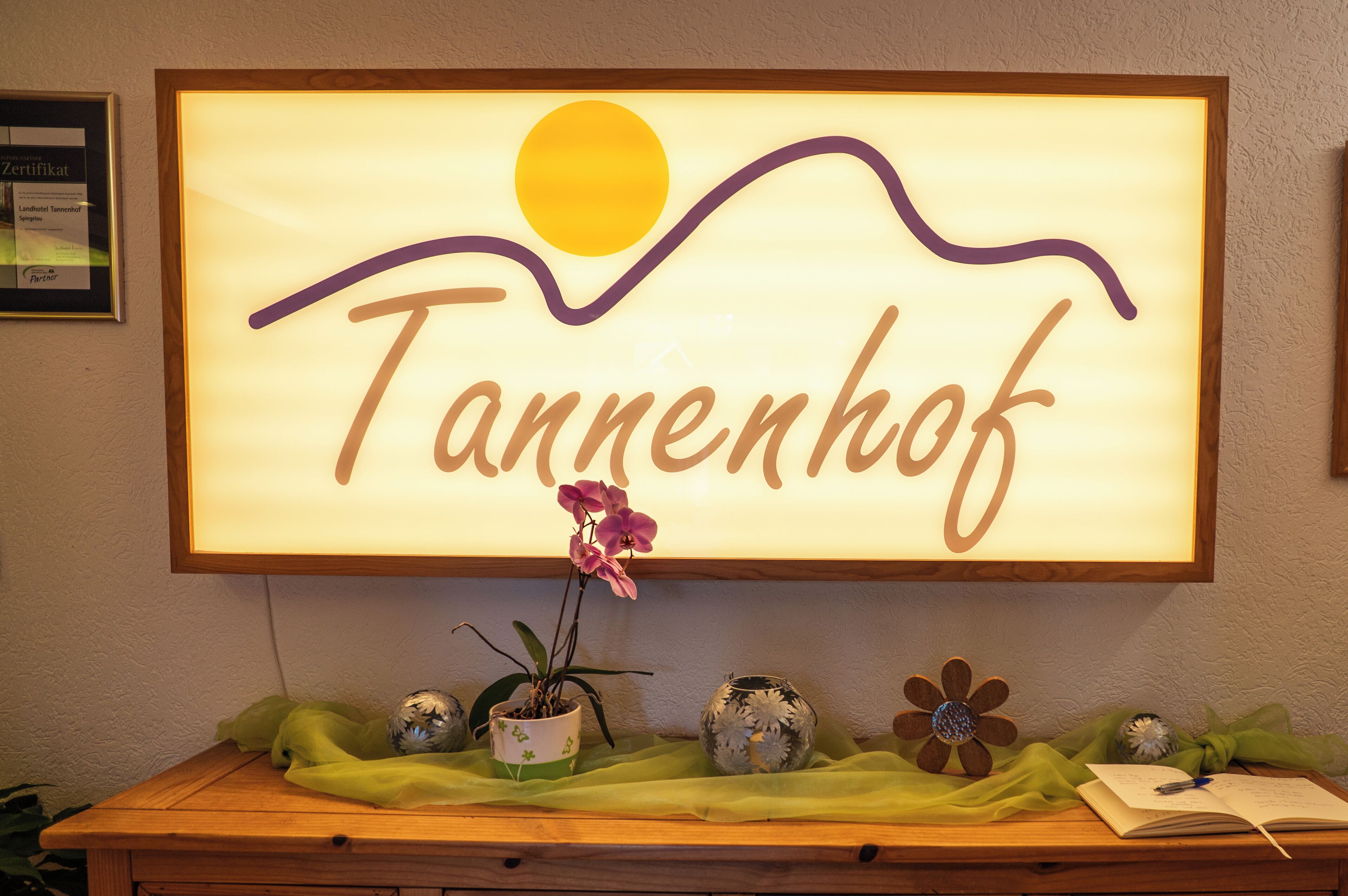 Ein gerahmtes Schild mit dem Wort "Tannenhof" und einer stützenden Sonne, darunter dekorative Elemente wie ein Blumentopf und eine blumenförmige Vase.