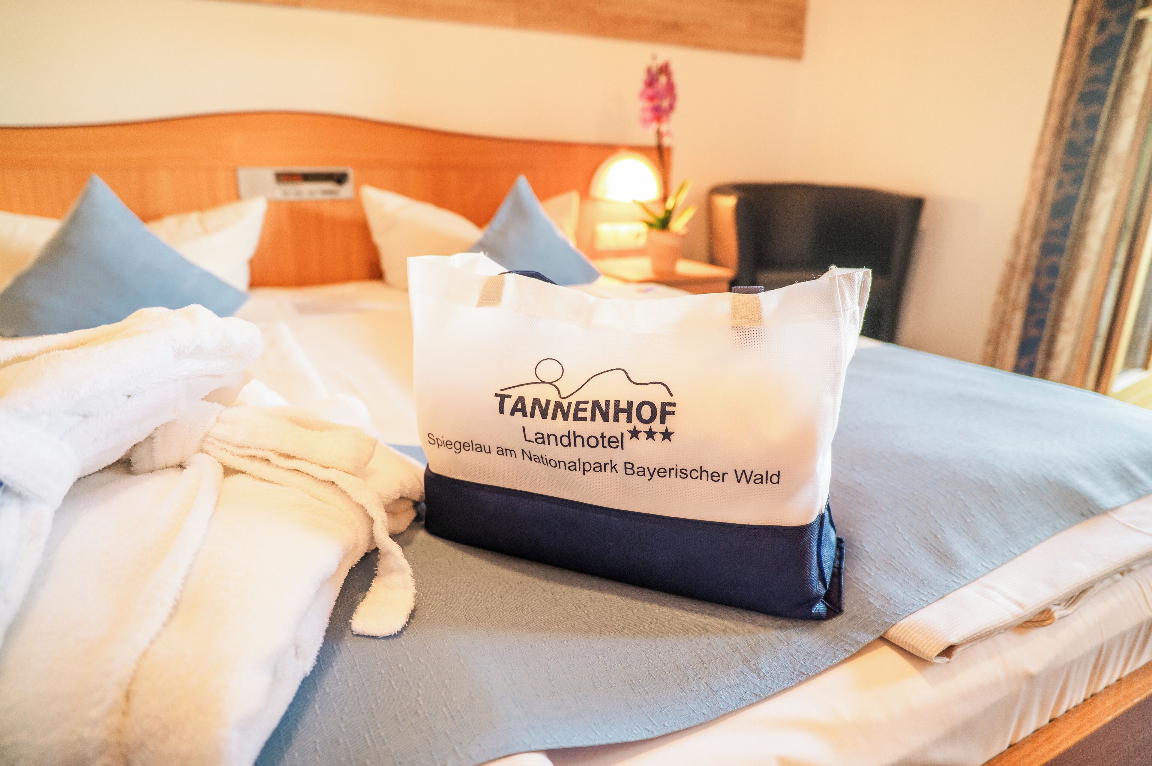 A hotel bed with a white pillow, a white towel, and a white bag with 'TANNENHOF Landhotel' text.