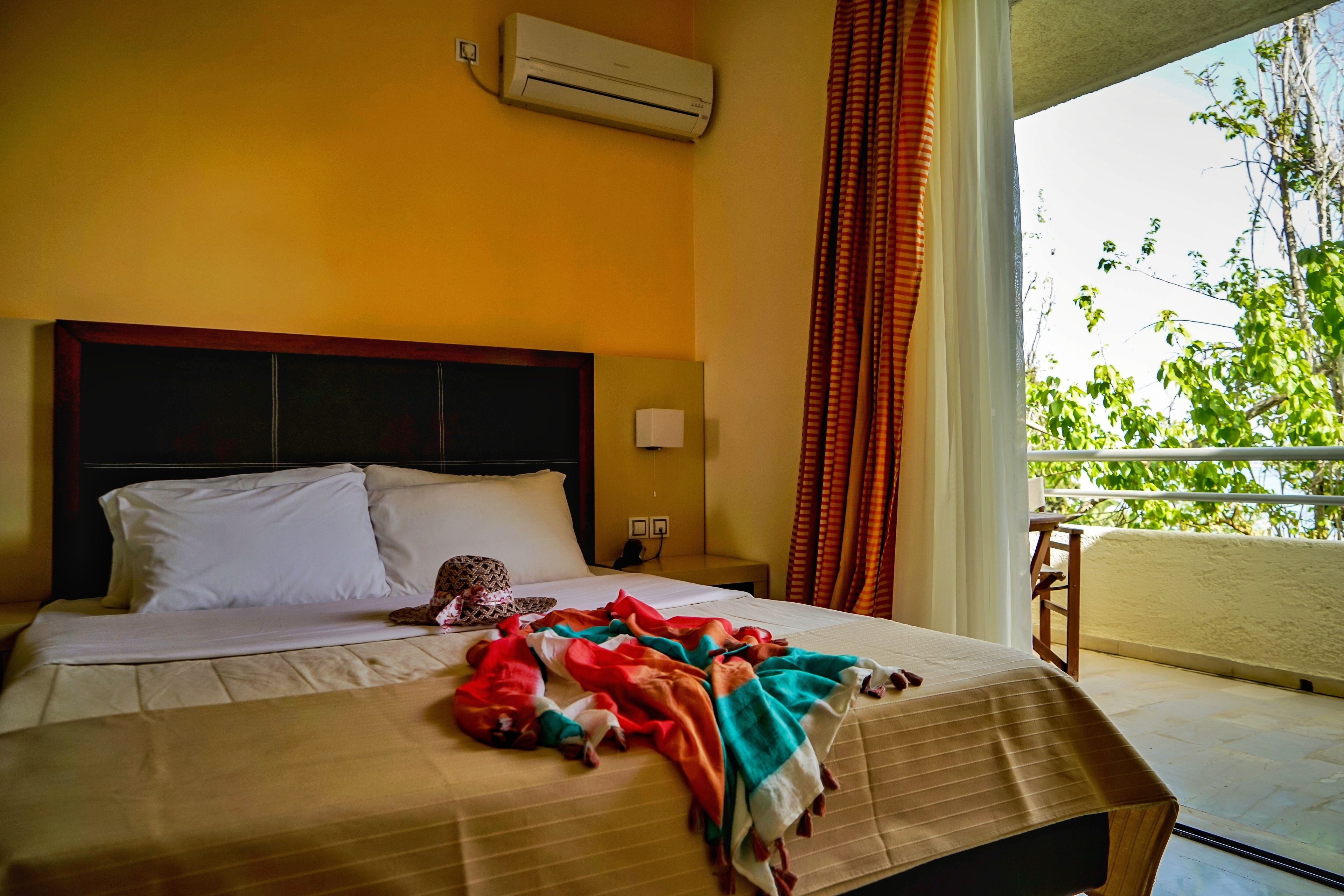 A hotel room with a bed, an air conditioner, a nightstand, a window with curtains, and a view of greenery outside.