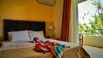 A hotel room with a bed, an air conditioner, a nightstand, a window with curtains, and a view of greenery outside.