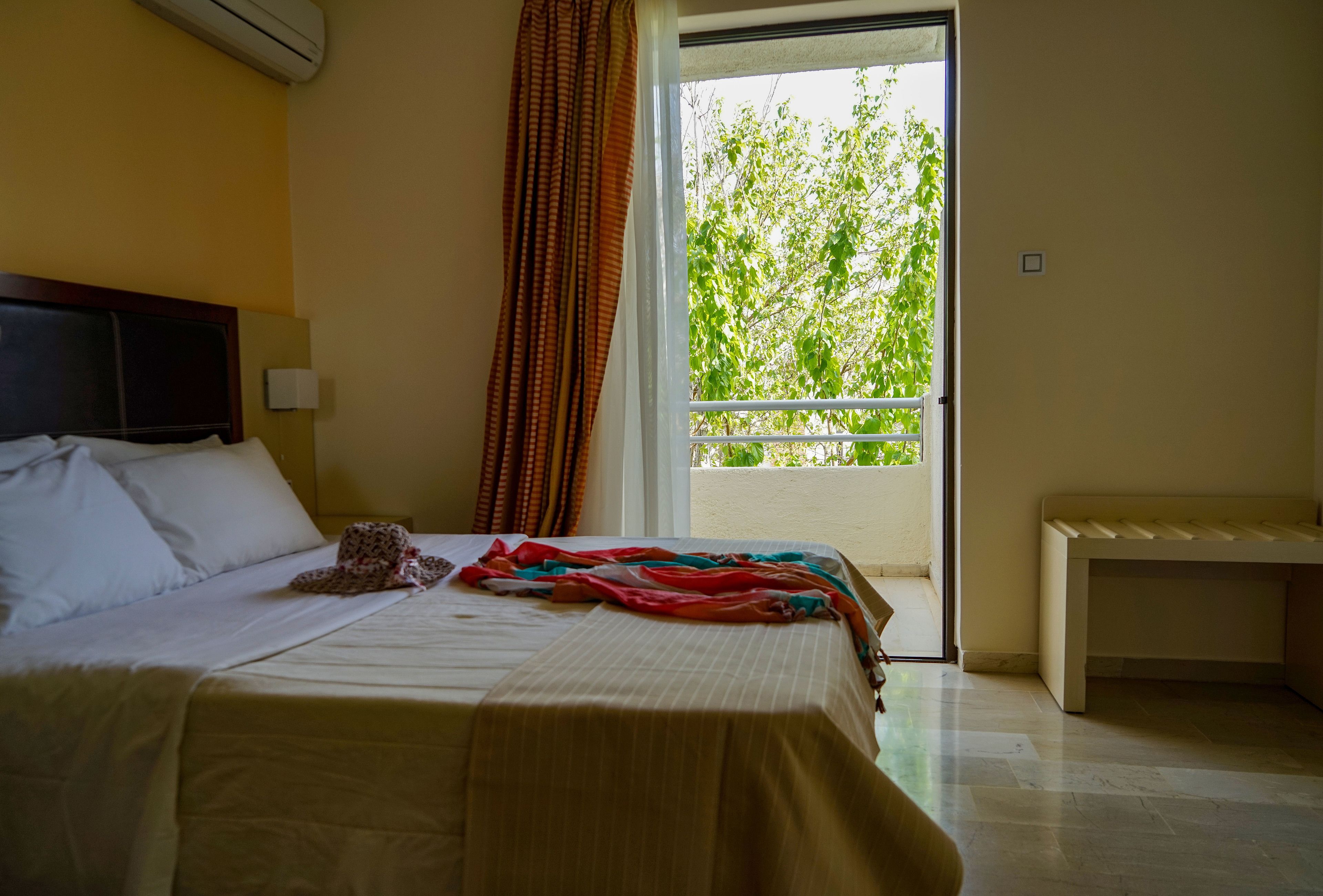Habitación de hotel con una cama, un banco, una ventana con vistas a la vegetación y una cortina.