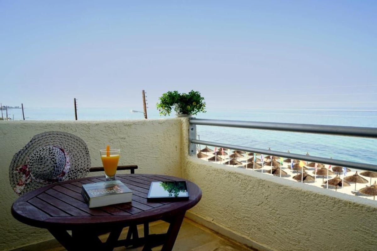 A round wooden table with a book, a glass of orange juice, and a straw. A woven hat is placed beside the table. A potted plant is on the balcony railing. The view includes a beach with umbrellas and the sea.