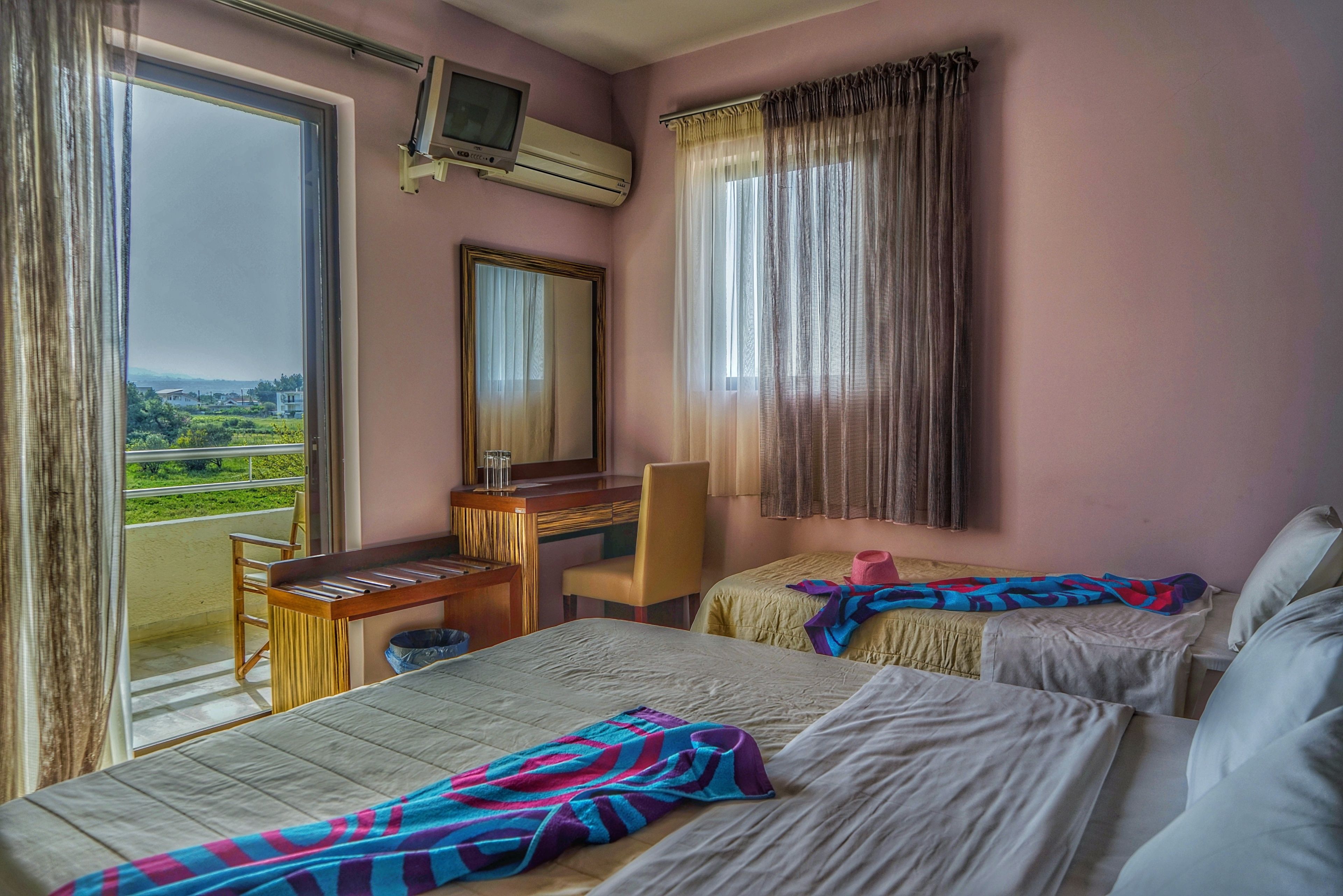 A hotel room with two beds, a desk, a chair, a TV, and a balcony with a view.