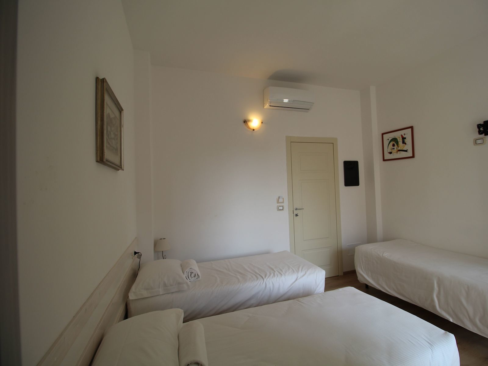 Una habitación con dos camas individuales, una puerta, un equipo de aire acondicionado y una lámpara fijada a la pared.