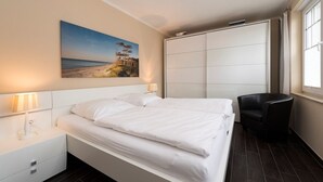 A bedroom with a bed, a nightstand, a chair, a wardrobe, and a painting on the wall. - Haus Ostseeblick (Göhren)