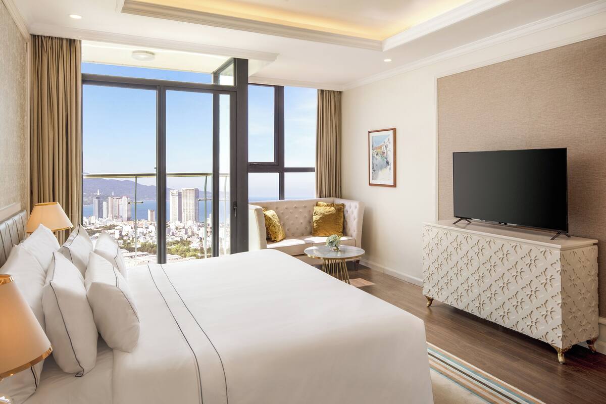 Ein Hotelzimmer mit einem großen Bett, einem Fernseher, einer Couch und Blick auf die Stadt.
