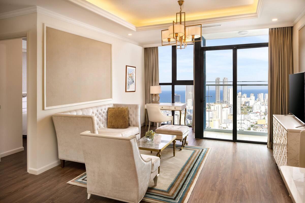 Ein modernes Wohnzimmer mit einer Couch, Sesseln, einem Couchtisch und einem Fernseher. Das Zimmer hat ein großes Fenster mit Blick auf die Stadt.