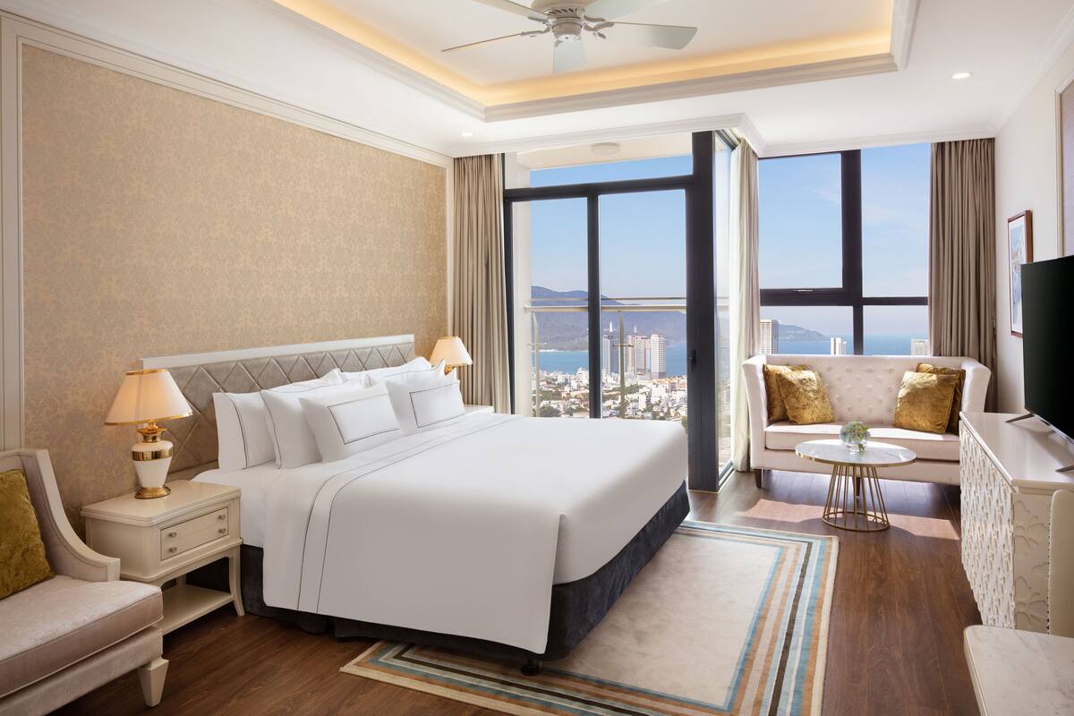 Ein modernes Hotelzimmer mit einem großen Bett, einer Sitzecke mit Sofa und Sesseln, einem kleinen runden Tisch und einem Fernseher. Das Zimmer verfügt über ein großes Fenster mit Blick auf die Stadt.
