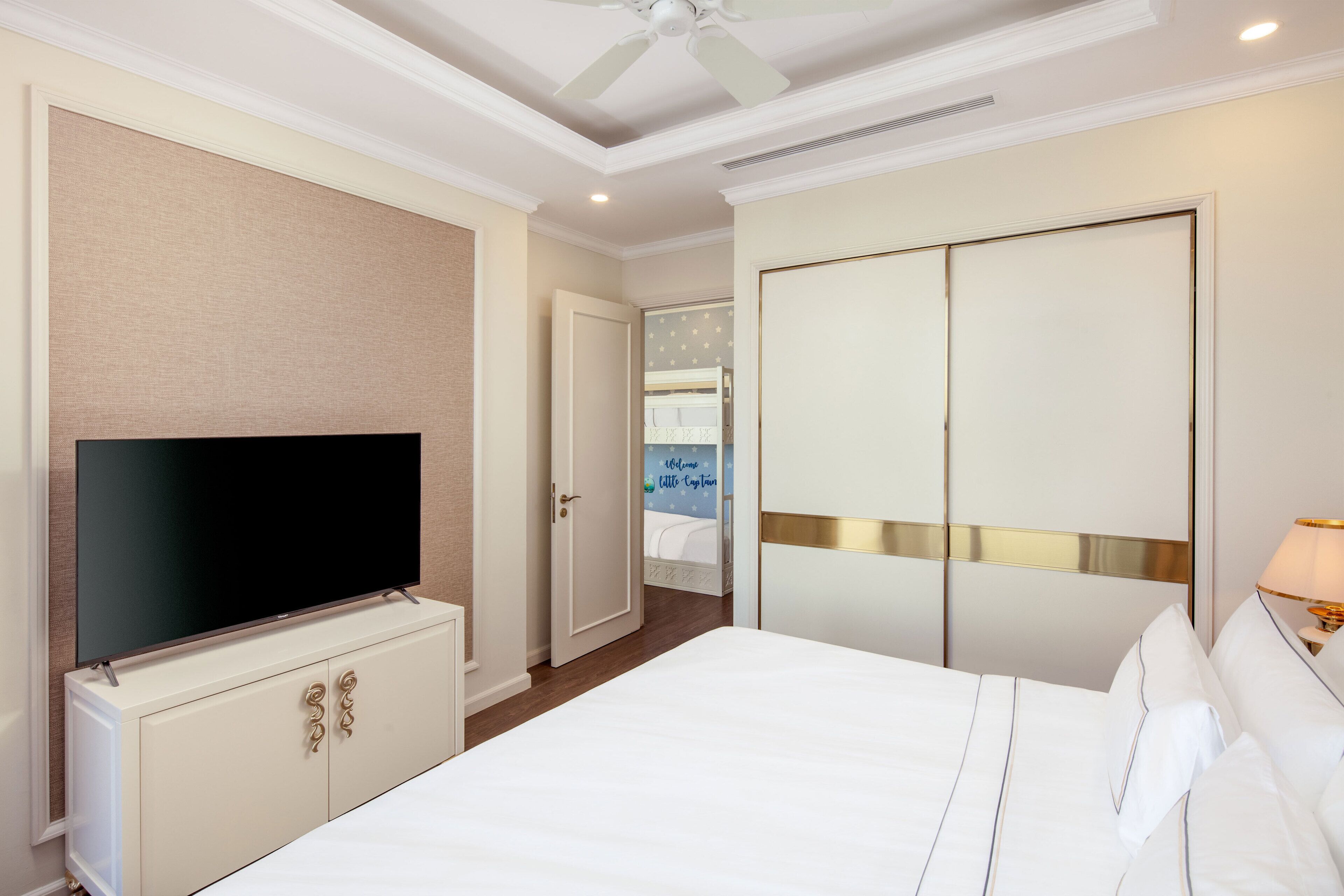 A hotel room with a flat-screen TV, a bed with white linens, a wardrobe with gold handles, and a ceiling fan.