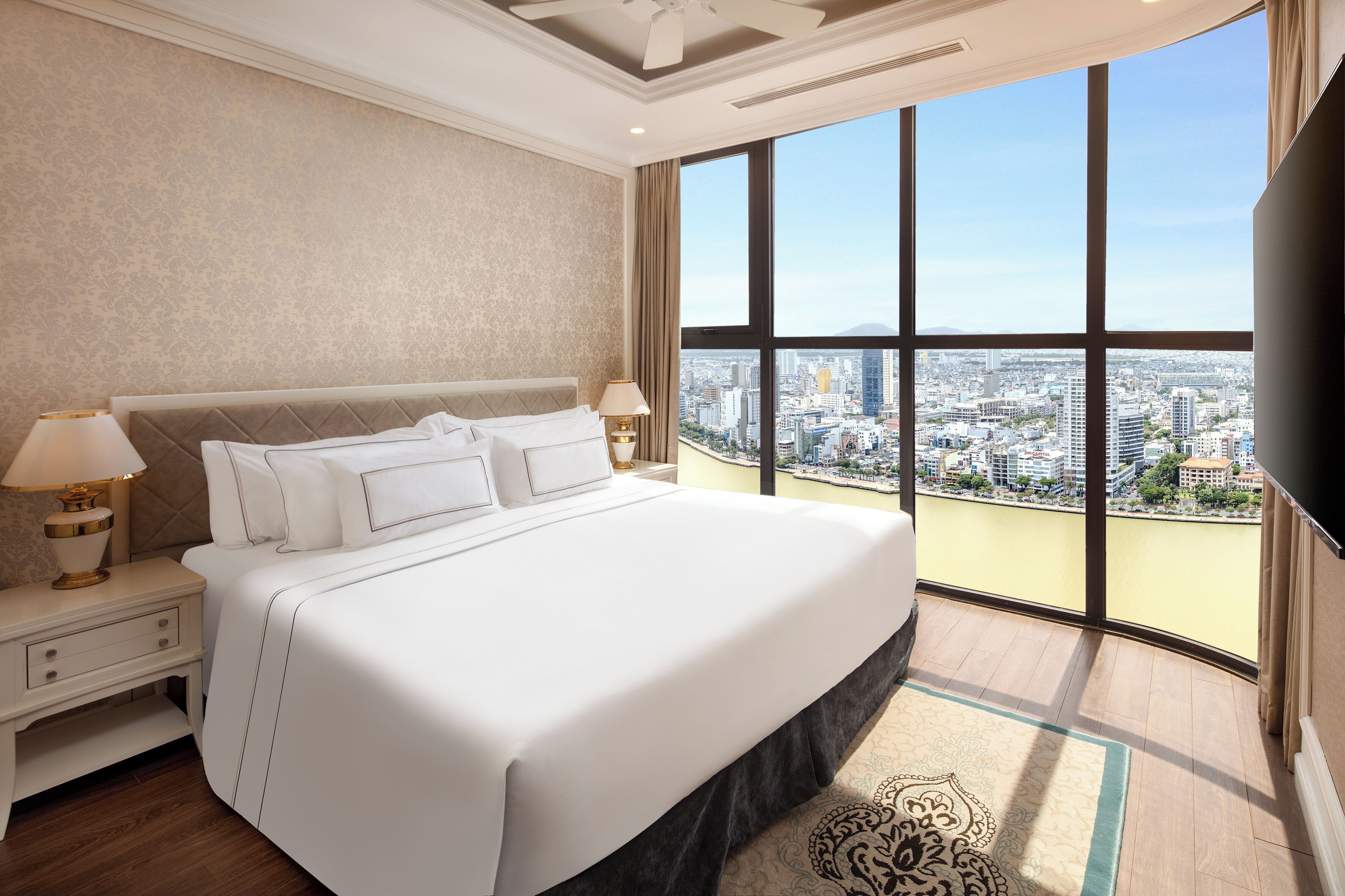 A hotel room with a large bed, two bedside tables with lamps, a TV, and a city view through large windows.