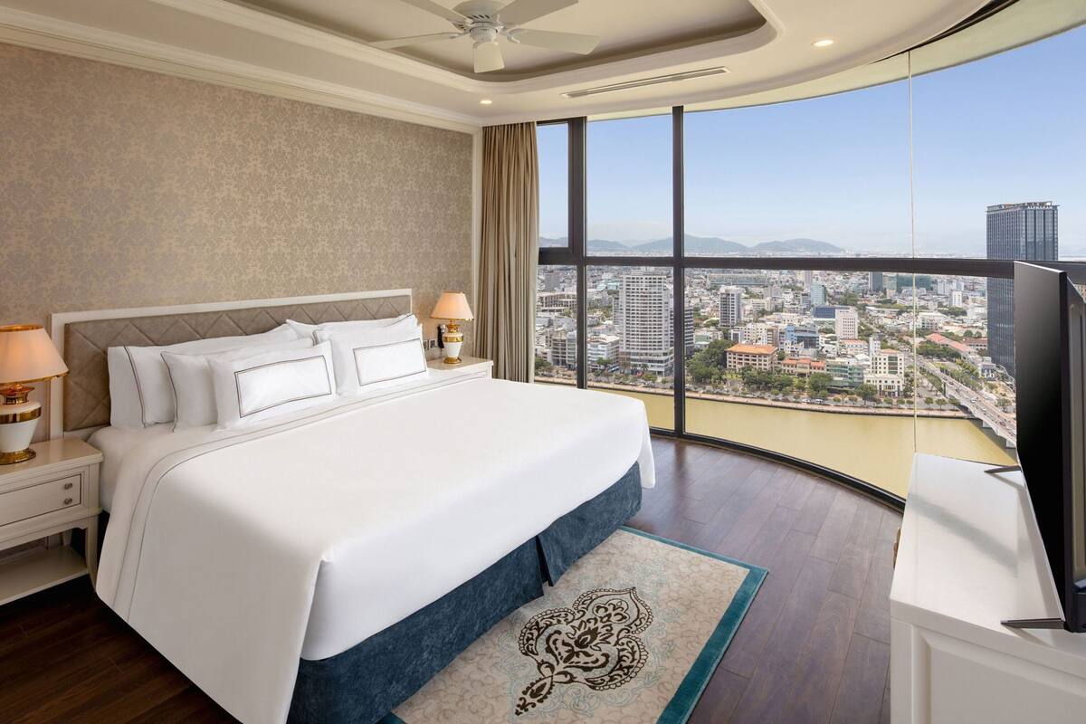 Ein Hotelzimmer mit einem großen Bett, Blick auf die Stadt, Deckenventilator und einem Fernseher.