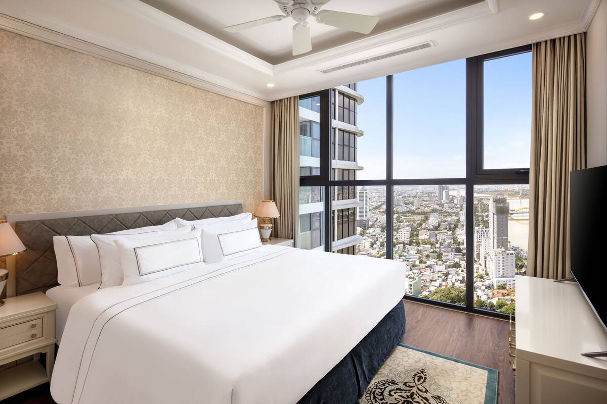 Ein Hotelzimmer mit einem großen Bett, einem Nachttisch, einem Fernseher und Blick auf die Stadt durch große Fenster.
