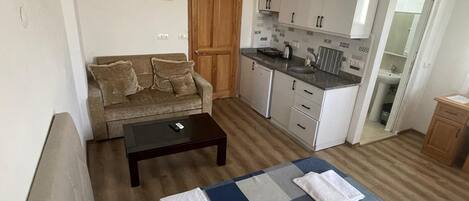 Un espacio habitable compacto con sofá, una mesa de centro, cama con colcha a cuadros, kitchenette y puerta de madera.