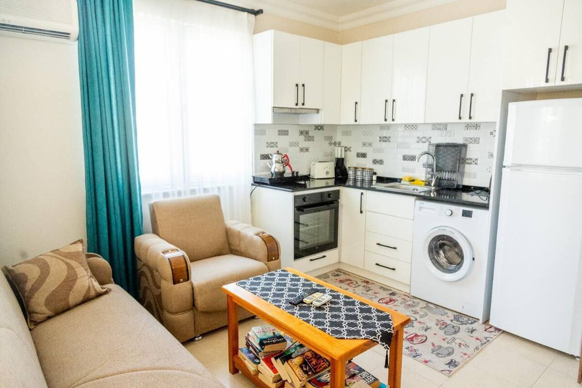 A compact living space with a sofa, a coffee table with books, a kitchen with a washing machine, and a window with curtains.