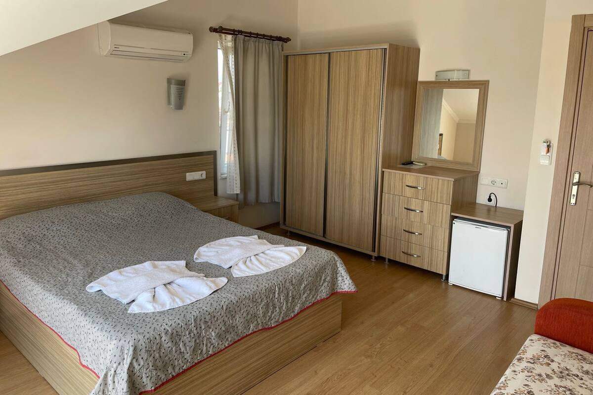 A hotel room with a bed, a wardrobe, a small desk with a mirror, and an air conditioning unit.