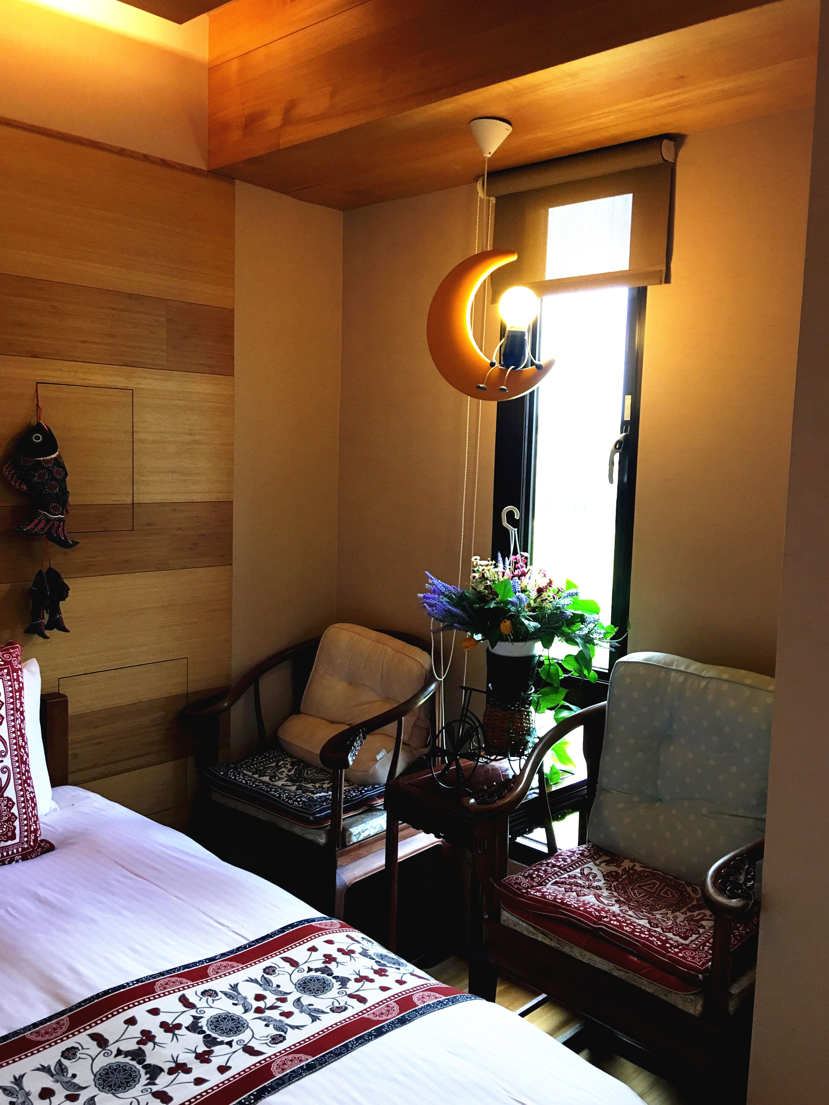 A bedroom with a bed, two chairs, a hanging light fixture, a window with a view, and a wooden wall.