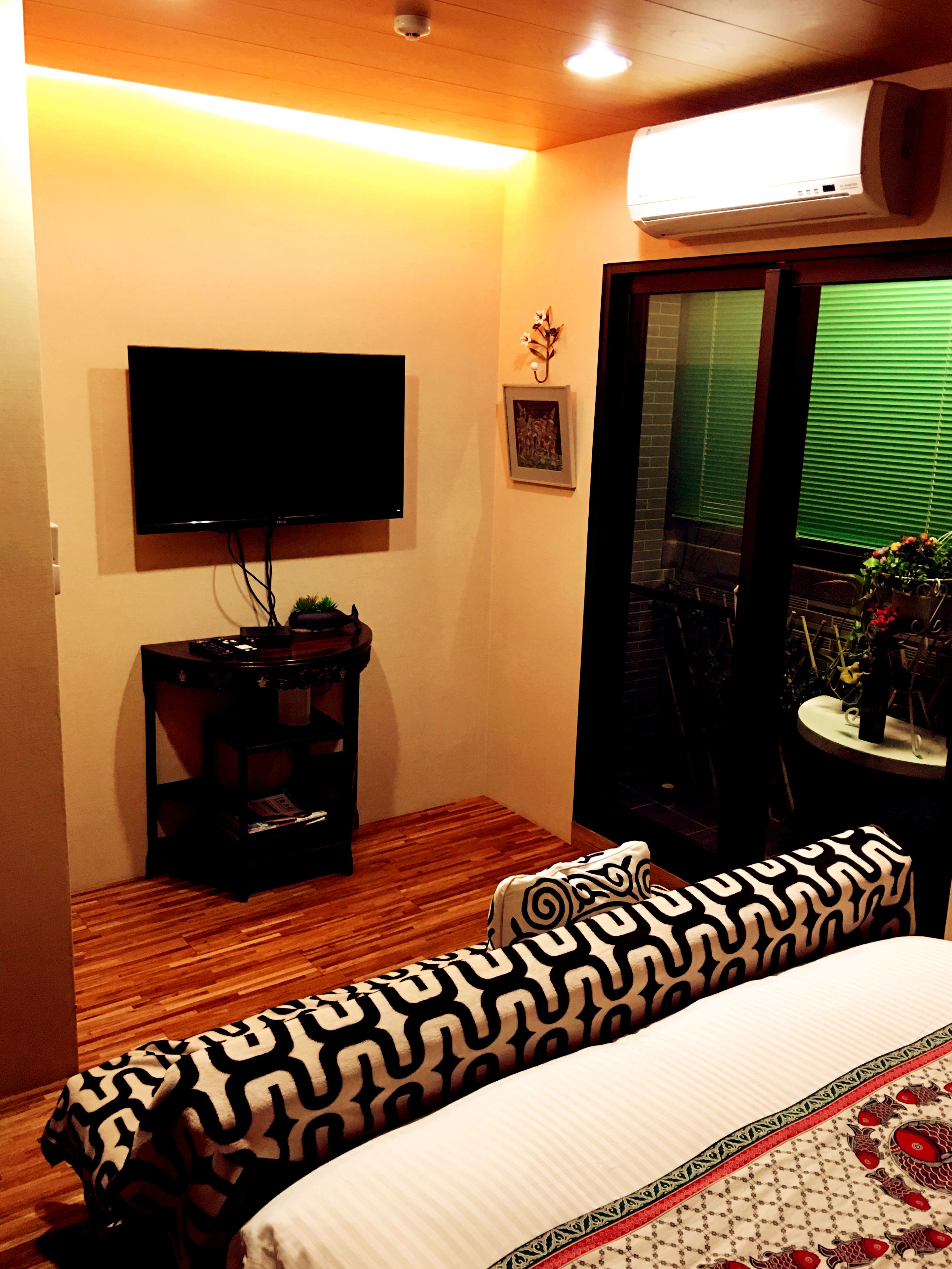A hotel room with a flat-screen TV mounted on the wall, a small table with a plant, a sofa with patterned cushions, and a window with blinds.