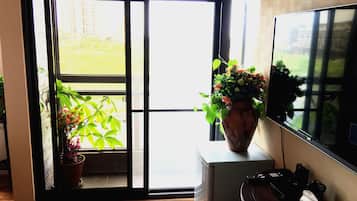 A room with a sliding glass door leading to a balcony, a large potted plant on the windowsill, and a flat-screen TV mounted on the wall.
