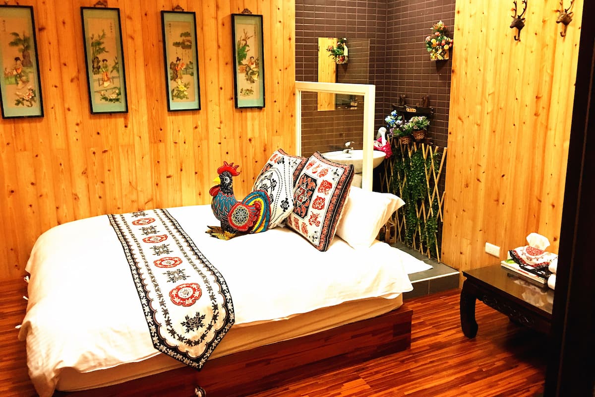 Une chambre à coucher bien rangée, avec un plancher en bois, un lit recouvert d’une literie à motifs, un coq décoratif et des œuvres d’art encadrées accrochées au mur.