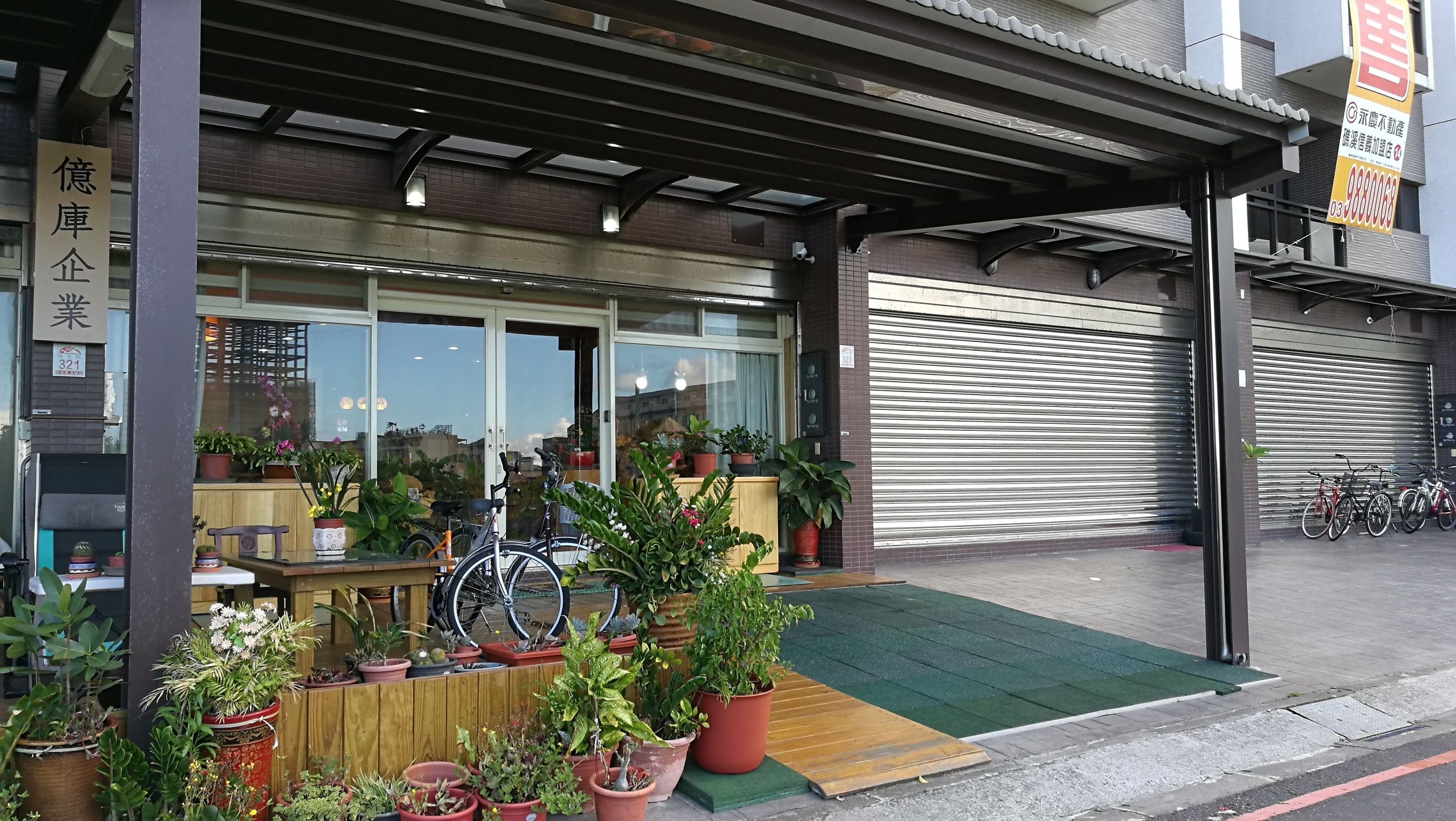 Un negozio con diverse piante in vaso esposte davanti, una bicicletta e un portone garage chiuso.