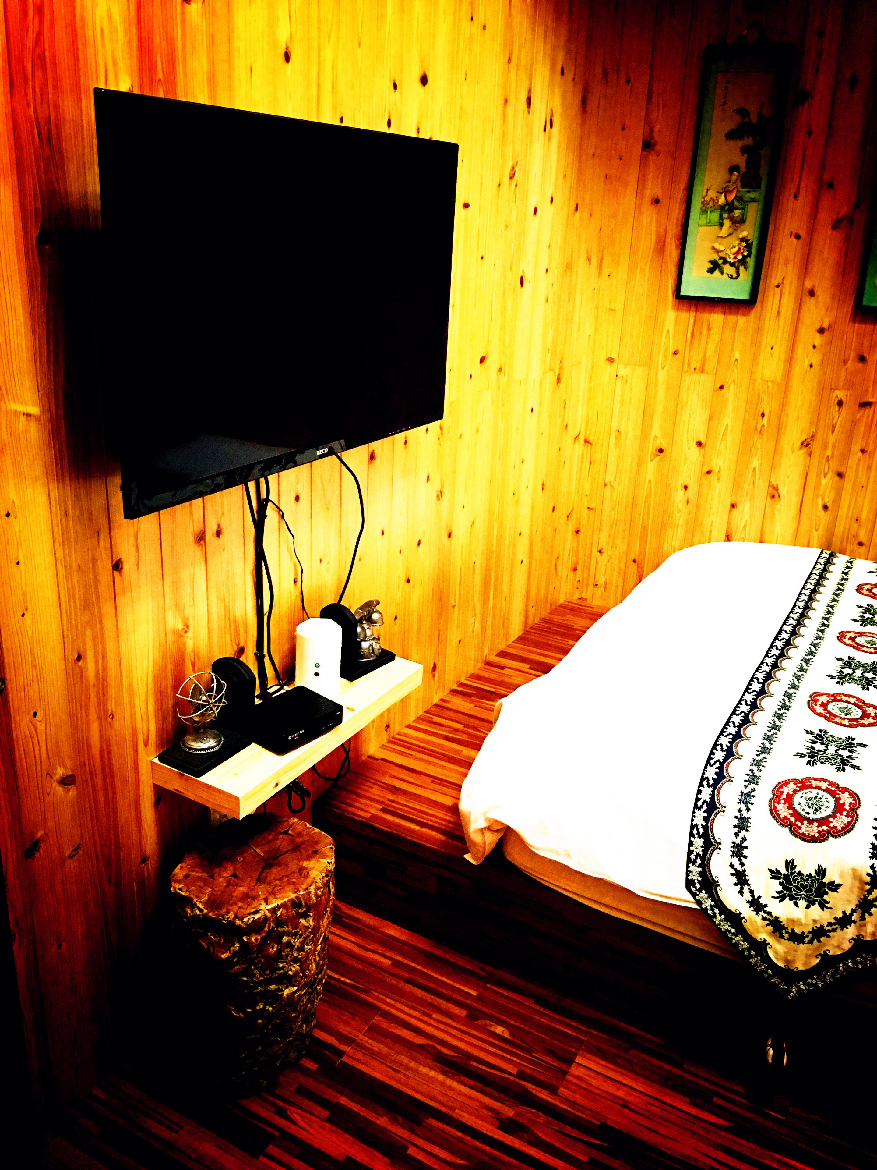 A room with a bed, a television mounted on a wooden wall, a small table with a lamp, and a decorative item on the floor.