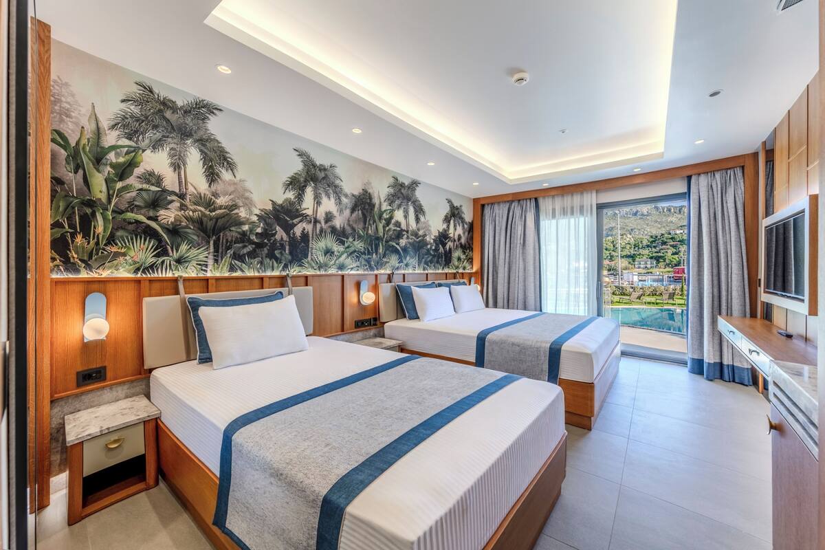 Quarto de hotel com duas camas, um grande mural de plantas tropicais, uma TV e uma varanda com vista para uma piscina e montanhas.