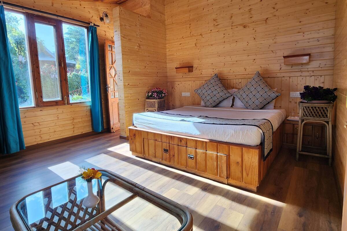 A wooden cabin room with a bed, a glass table, and a wicker chair.