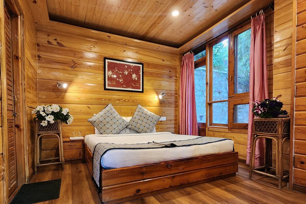 A wooden cabin bedroom with a bed, a nightstand, a framed picture, and a window with curtains.