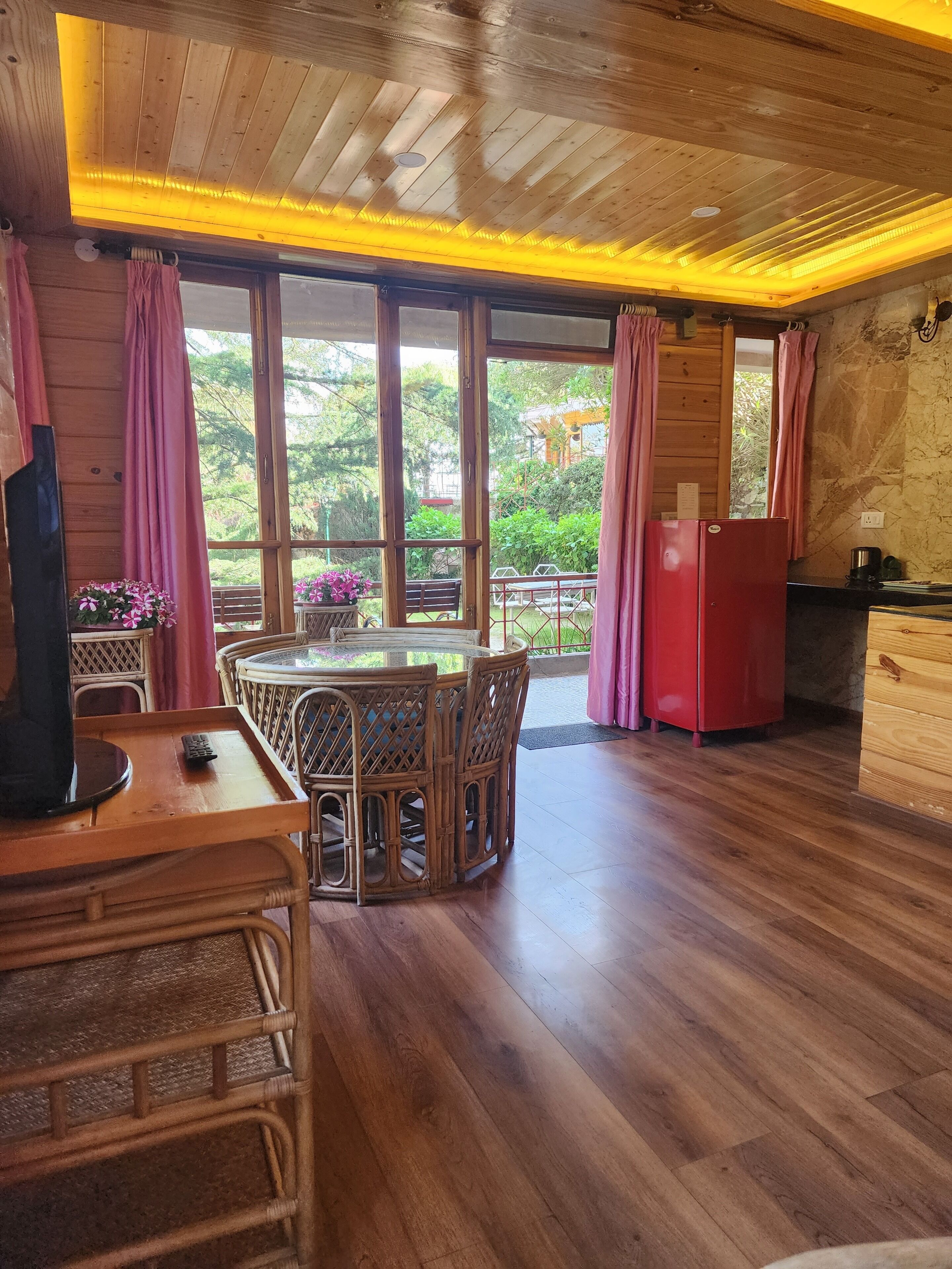 A wooden interior with a dining area, a red refrigerator, and large windows.