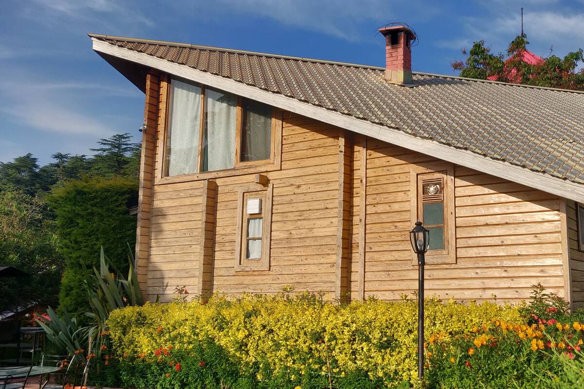 A wooden cabin with a sloped roof, surrounded by a garden with various flowers.
