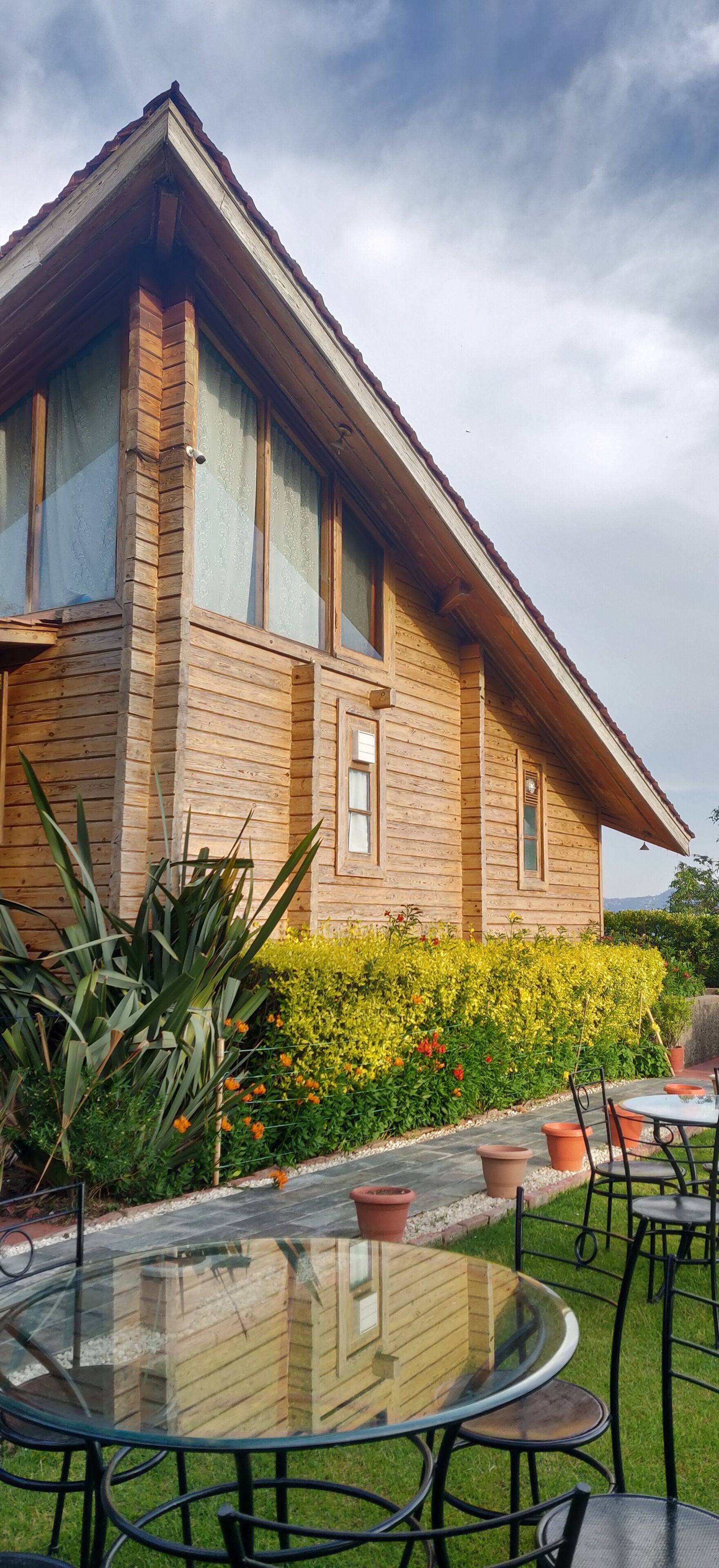 Una casa de madera con techo inclinado, ventanas amplias y una mesa de vidrio con sillas al frente.