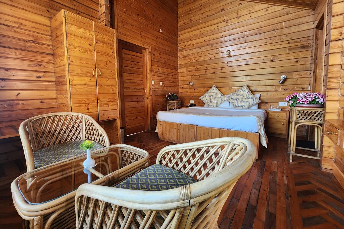 A wooden cabin room with a bed, two wicker chairs, a small table, and a nightstand.