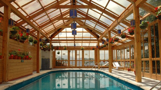 Una piscina interior con vigas de madera y paredes de cristal, rodeada de plantas en macetas y tumbonas.